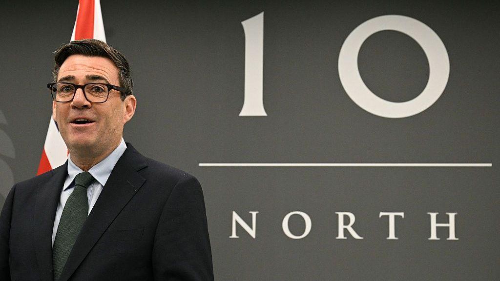 Andy Burnham stands in front of a Number 10 North sign while smiling.
