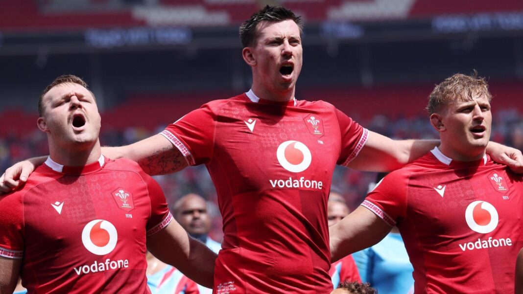 Adam Beard in the centre has his arms around the shoulders of Dewi Lake and Jac Morgan as the three Wales players in red jerseys sing the national anthem before a game.
