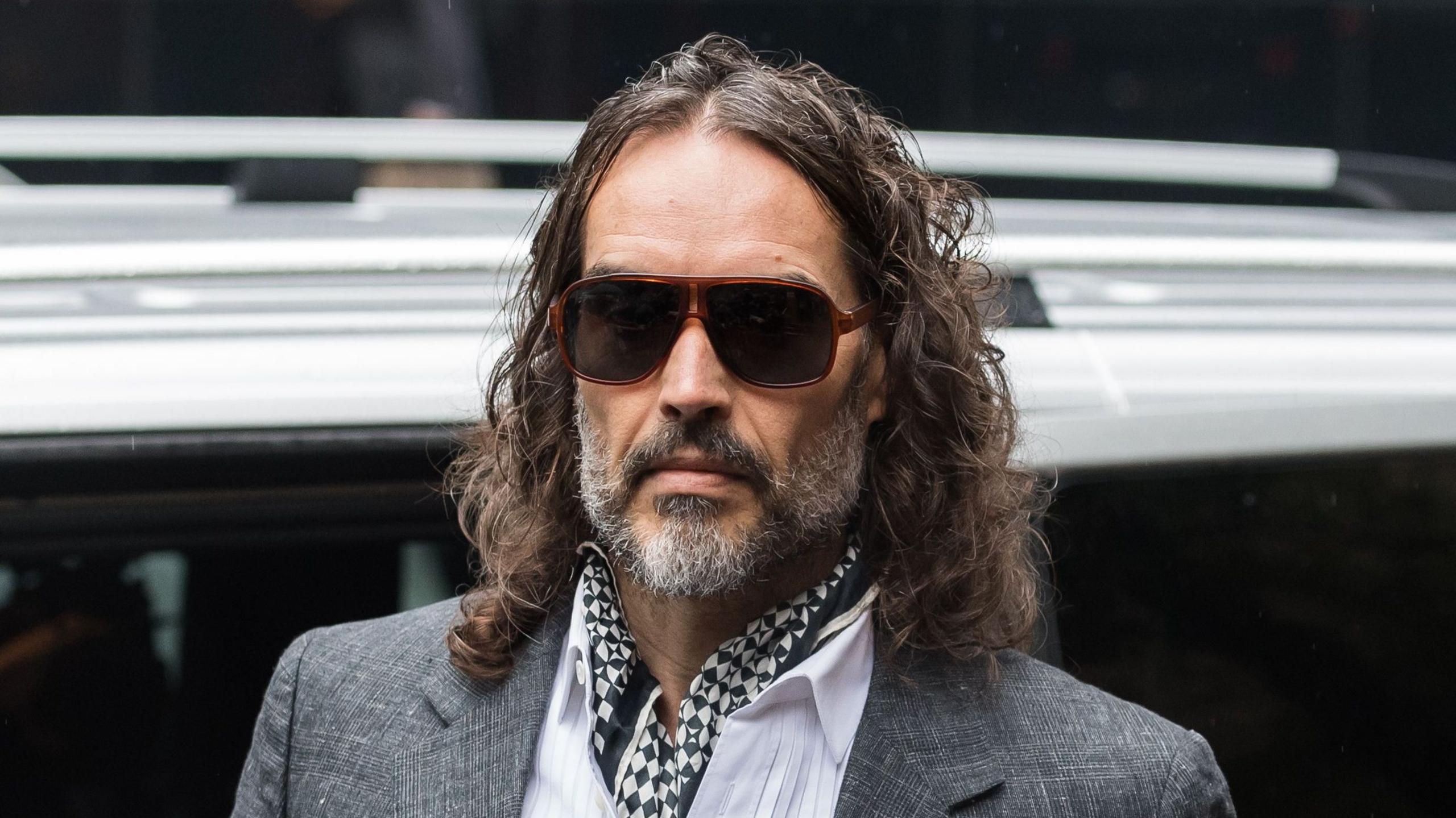 Russell Brand arriving at court on Monday 26 October - he is wearing a grey suit jacket, white shirt and black and white cravat. He is also wearing sunglasses.