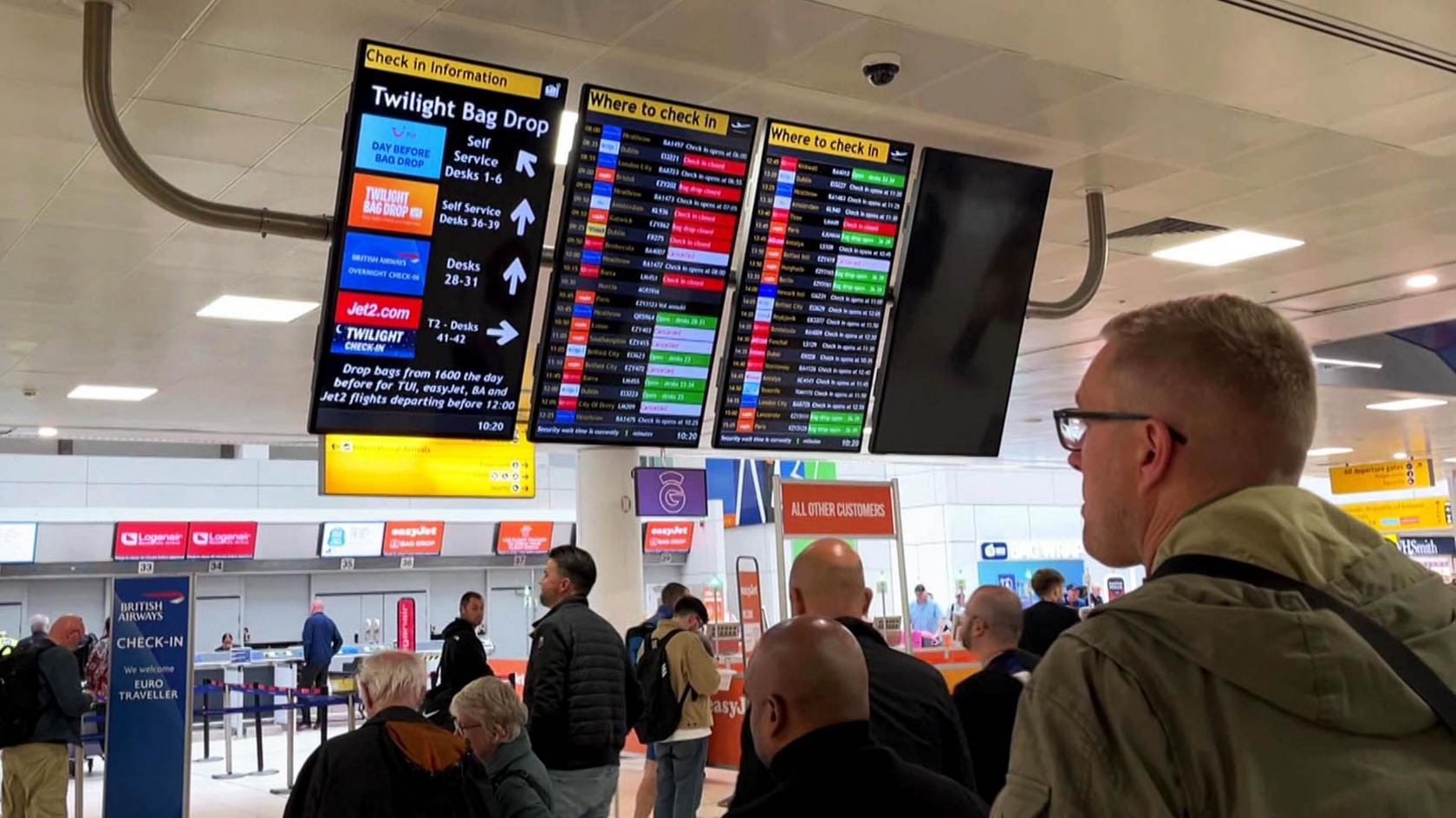 Shot from behind several passengers waiting at departure boards for information at Glasgow airport.