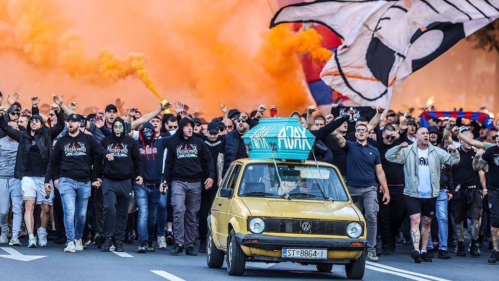 A large group of fans walk behind a small yellow car with a blue coffin on the roof with 'Ajax' written on it. The fans are waving a big flag and letting off orange flares.