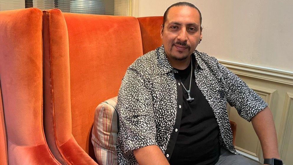 a man in a black t-shirt and open black and white animal print shirt is sat on an orange velvet chair, looking directly at the camera with a calm smile