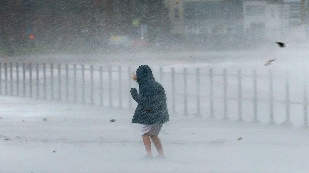 A person wearing shorts and a waterproof cowers as waves splash over the promenade spraying water everywhere