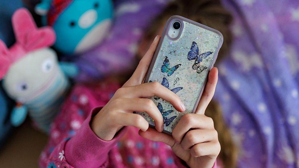 A young girl with long brown hair lying on her bed with a purple and white duvet, looking at her phone over her face, which has butterfly stickers on the back. There are two out-of-focus toys next to her in blue and white, and she is wearing a pink jumper with white and blue flowers on it.