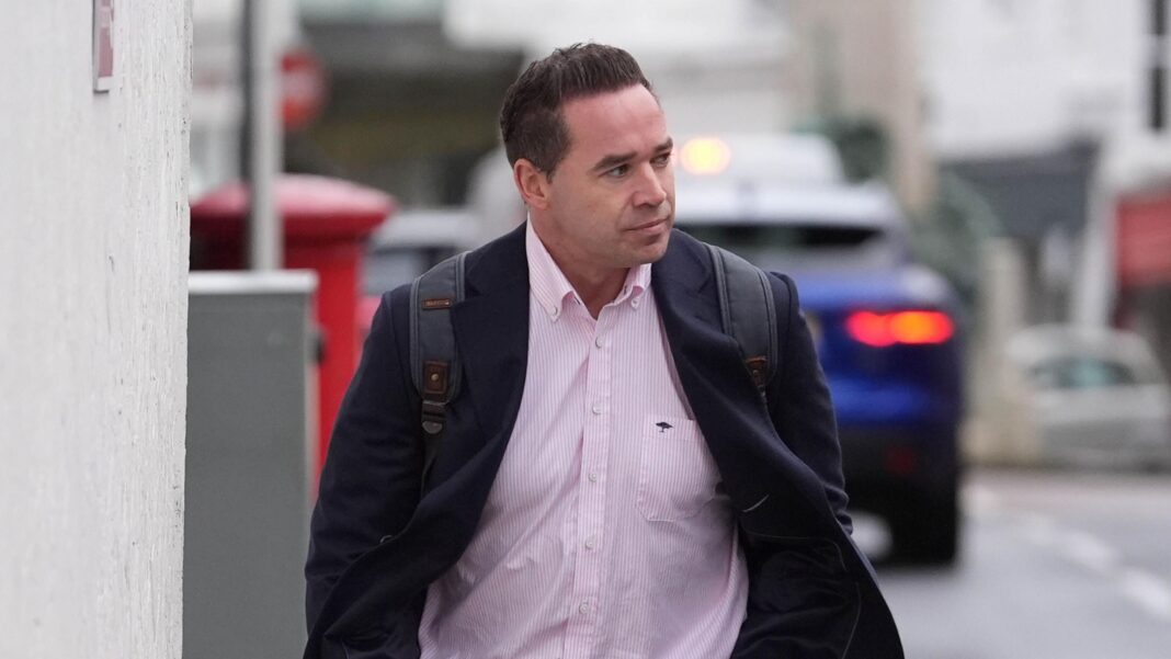 Kieran Hayler walking along a street, he is wearing a pale pink striped shirt with a dark jacket and a rucksack. He has short dark hair, and is looking away from the camera.