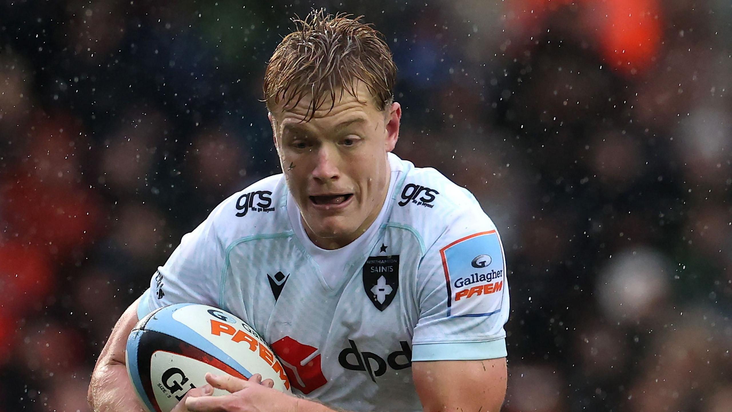 Fin Smith in a white Northampton Saints jersey during a match. He is carrying a rugby ball as rain falls around him.