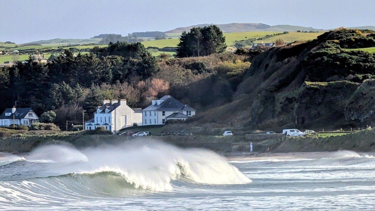Big waves crash over to create surf water in the sea, with houses, trees and hills behind