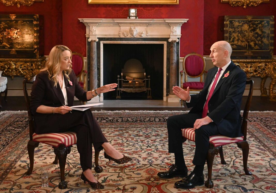 Laura Kuenssberg sat in a chair on the left opposite Chancellor John Healey on the right. They are in conversation and using hand gestures. Kuenssberg wears a dark brown coloured suit and Healey is wearing a black suit. They are sitting in an ornate room in front of a fire place.