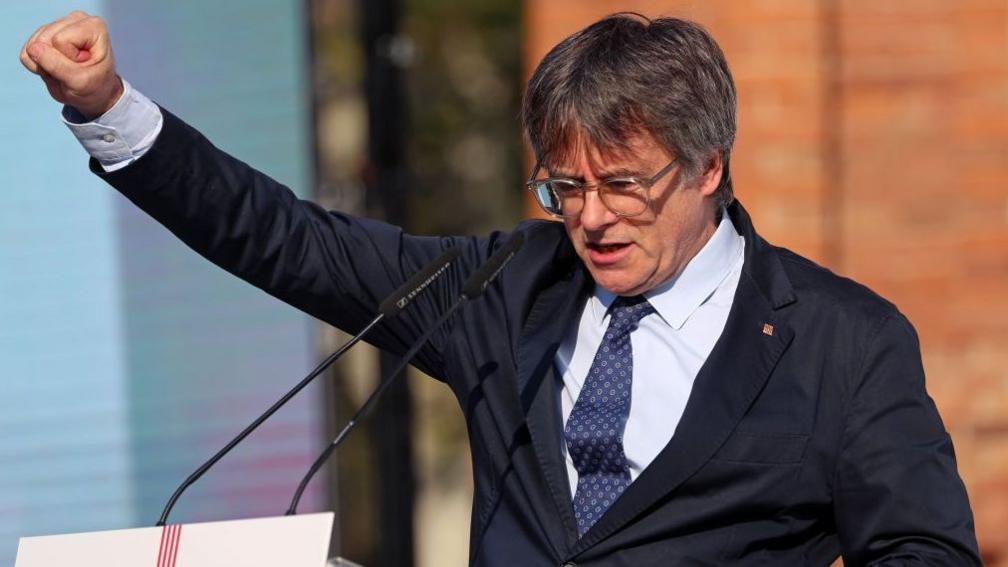Carles Puigdemont pictured in Barcelona in August 2024. He is a middle aged man with grey-brown hair. He wears large round glasses, a dark blue suit jacket, white shirt and patterned dark blue tie. He is stood in front of a podium at an outdoor rally, and has his right fist raised.