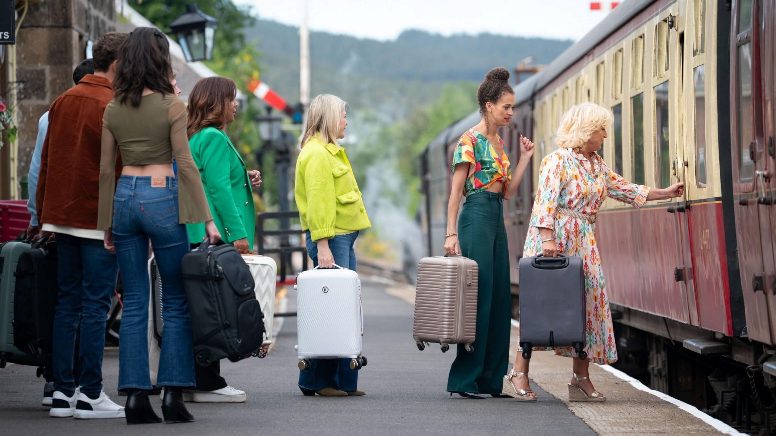 The Traitors contestants stand in a line holding suitcase on a railway platform. The contestant at the front of the line, a woman wearing a brightly patterned dress is reaching out to open a door of cream and claret coloured carriage.