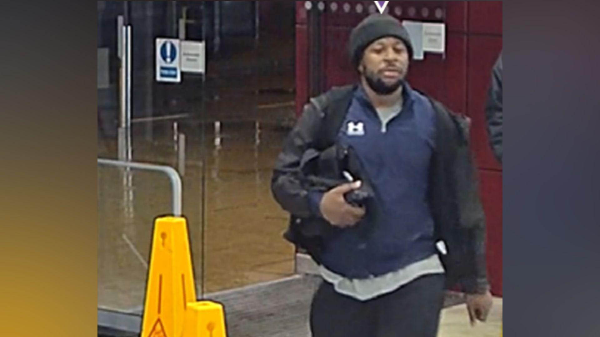A man in dark clothing and black woolly hat carries a small black bag under his right arm. He is outside glass doors at a railway station and is walking past yellow cones that say "CAUTION WET FLOOR" on them.