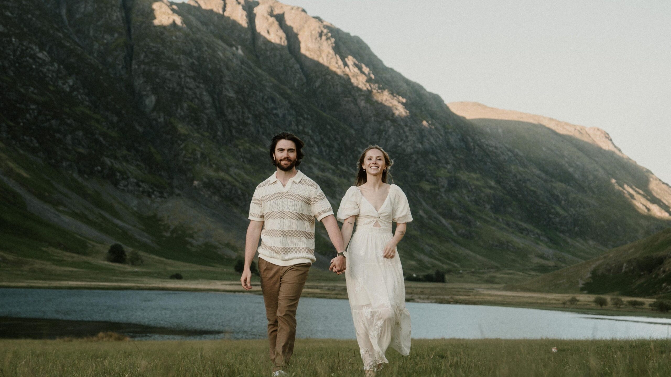 The couple stand in front of a small body of water, with a mountain valley behind them. They hold hands while smiling down the camera, Savannah where a white dress while Hayden where brown trousers and brown striped polo.