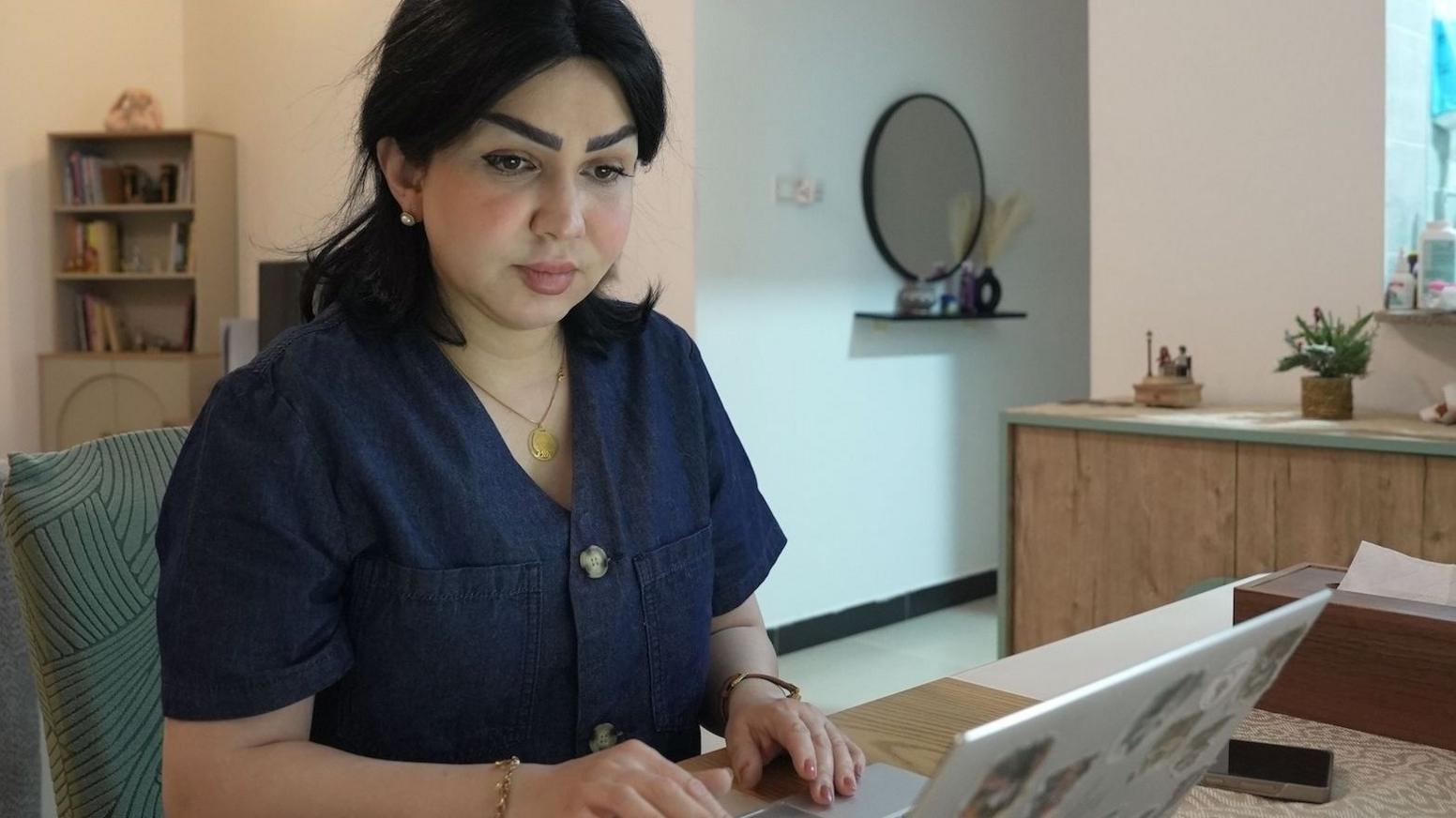 Sarah Jassim sitting at a desk typing on a laptop in a room with a bookshelf, flowers and a mirror in the background. She has dark, mid-length hair and is wearing a short-sleeved blue top.
