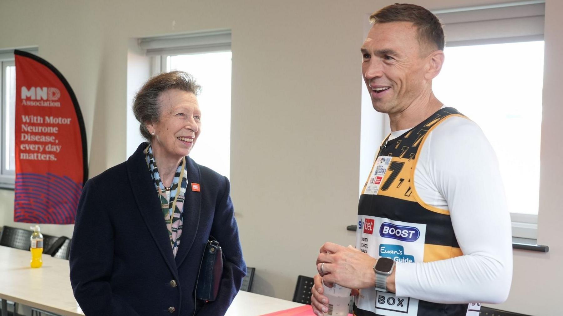 Kevin Sinfield in a black-and-yellow running vest holds a water bottle while chatting with Princess Anne wearing a dark coat and patterned scarf. An MND Association banner stands beside the windows.