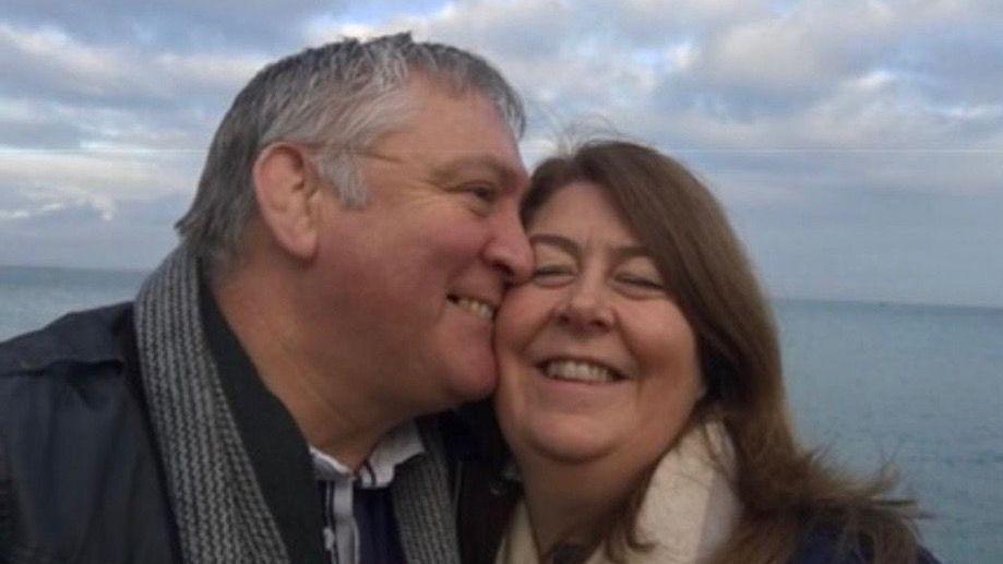 A selfie of John kissing Louisa on the cheek they are both wrapped up warm in coats and scarves and smiling as the wind blows their hair with a blue cloudy sky and sea behind them