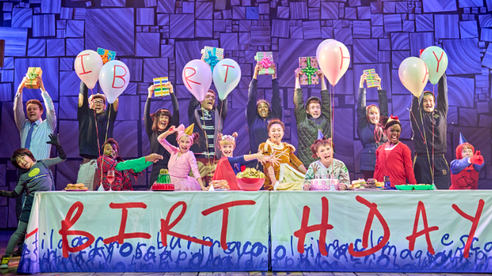 8 child actors are on stage in different fancy dress costumes, they're celebrating a birthday with balloons, food and presents. They are all stood in a different pose during the scene with the actors playing their parents stood behind them holding presents in the air.