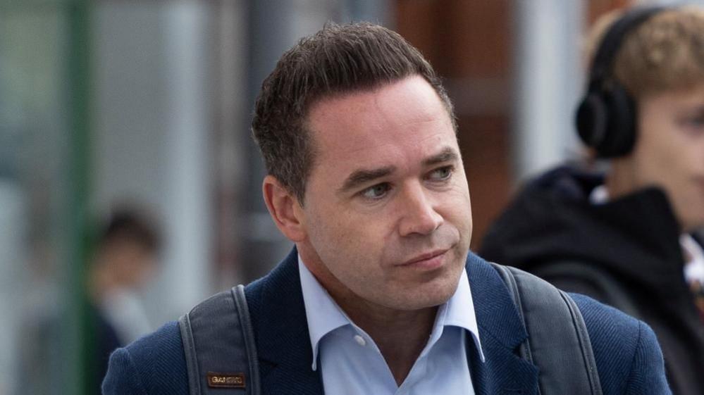 A man, Kieran Hayler, wearing a blue suit, white shirt and a backpack. He is looking to his left.
