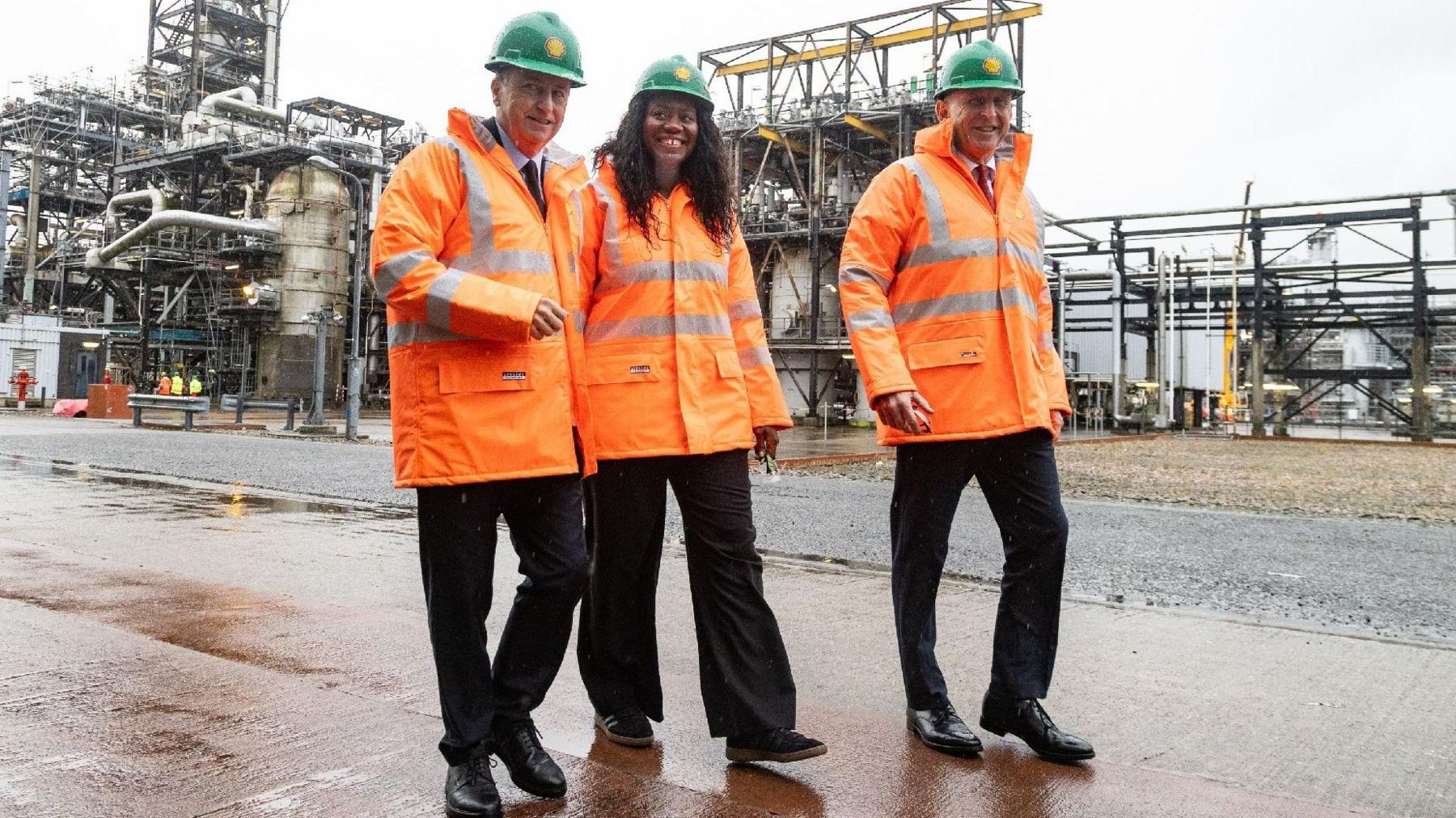 Scottish Secretary Douglas Alexander, Energy Secretary Miatta Fahnbulleh and Chancellor John Healey during a visit to Shell UK's St Fergus Gas Terminal in Peterhead, Aberdeenshire. They are wearing orange high-vis jackets and green hard hats, as they walk past gas terminal infrastructure in the rain.