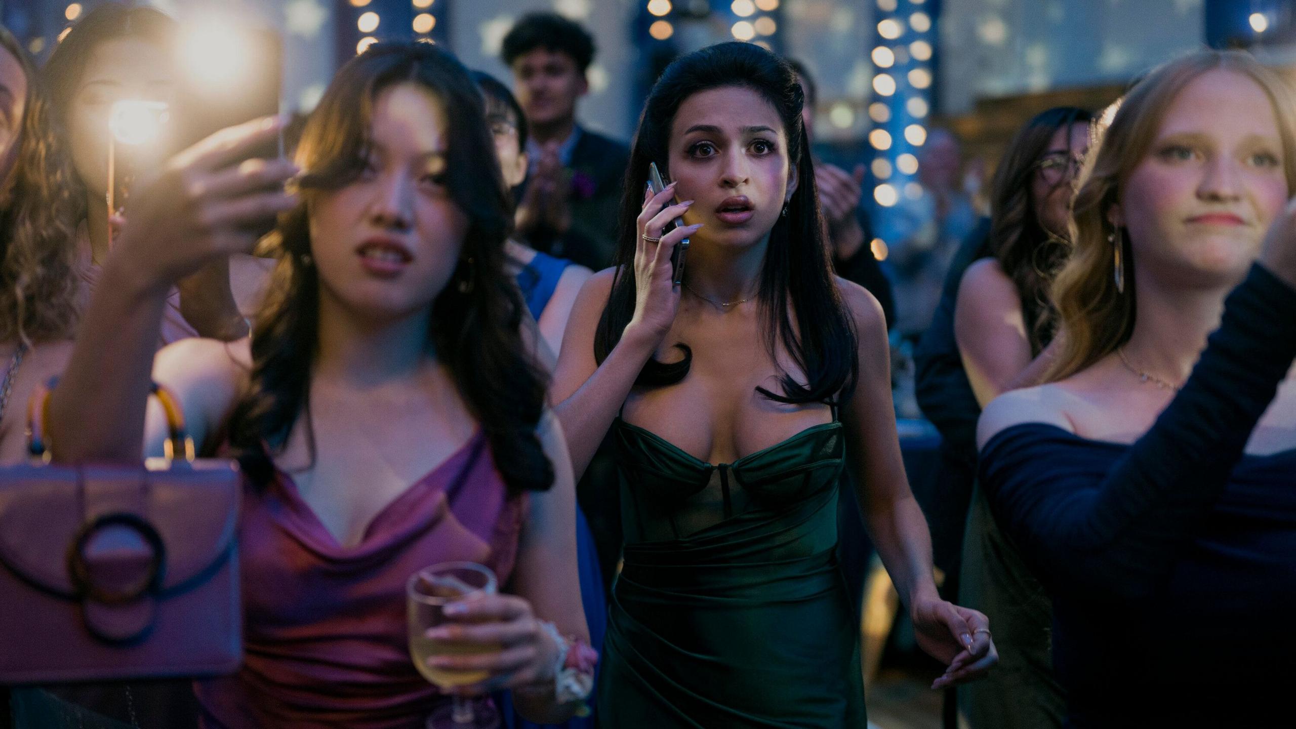 Still from Carrie shows Tina, wearing a green prom dress, standing in the middle of a crowd of high schoolers at prom. They are all holding their phones, filming something in front of them. Tina, however, appears to be making a panicked phone call, with her mobile phone pressed to her ear and a look of concern on her face.