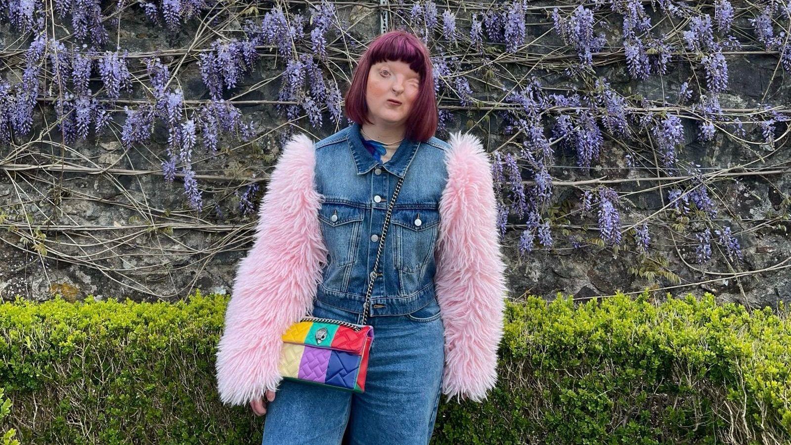 Kirsty with a purple bob is standing in front of a wall of purple flowers, she has a neutral expression. She's wearing a denim outfit with pink fluffy sleeves and a bright rainbow-coloured handbag.