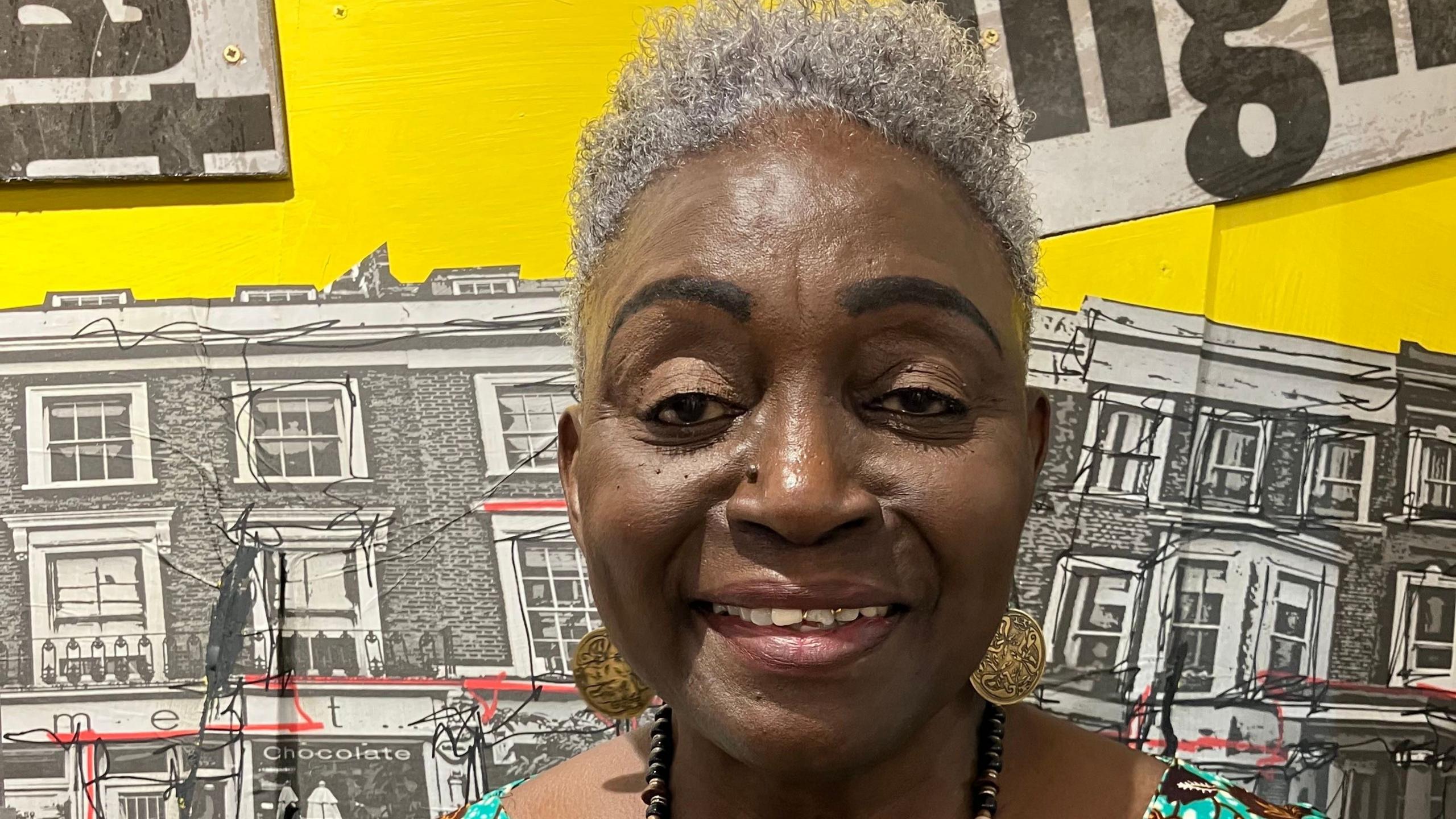 A smiling Allyson wears gold earrings and a beaded necklace. Behind her part of the exhibition can be seen showing yellow paint and drawings of Notting Hill buildings.