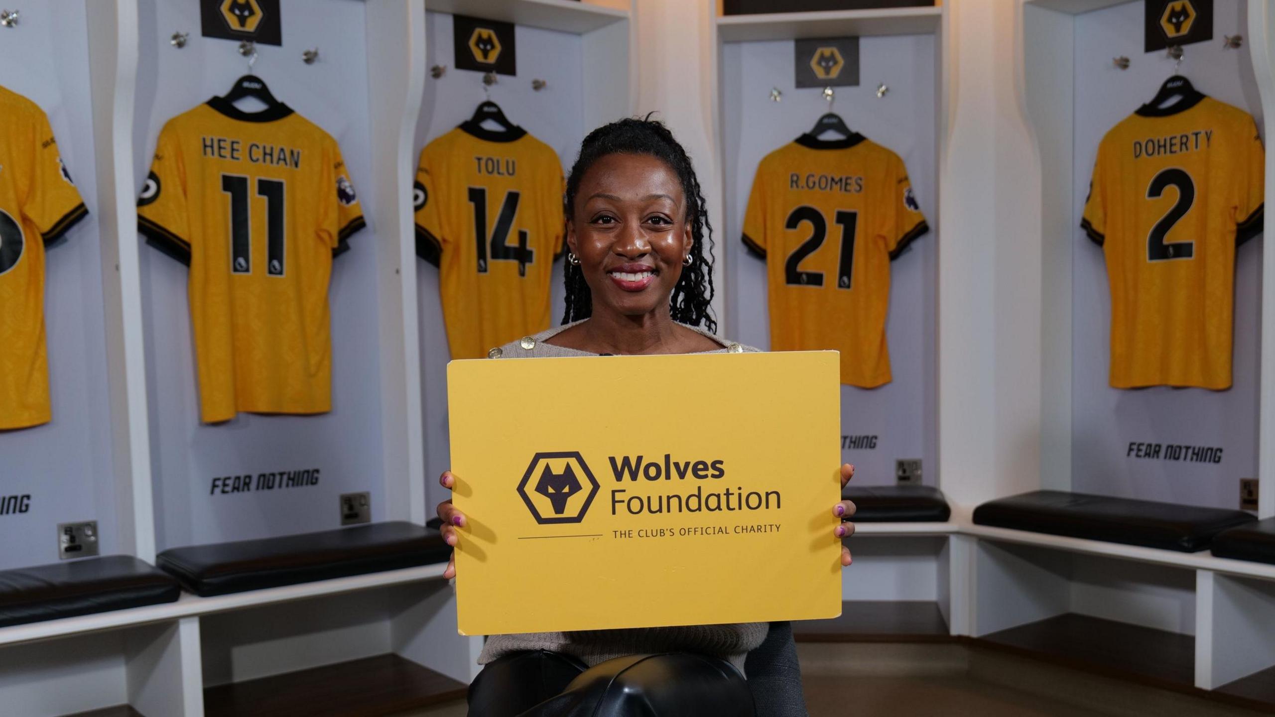 Beverley Knight, a woman with her hair in a ponytail is said in a football changing room surrounded by Wolves FC shirts and holding a yellow board which reads Wolves Foundation with a wolf logo