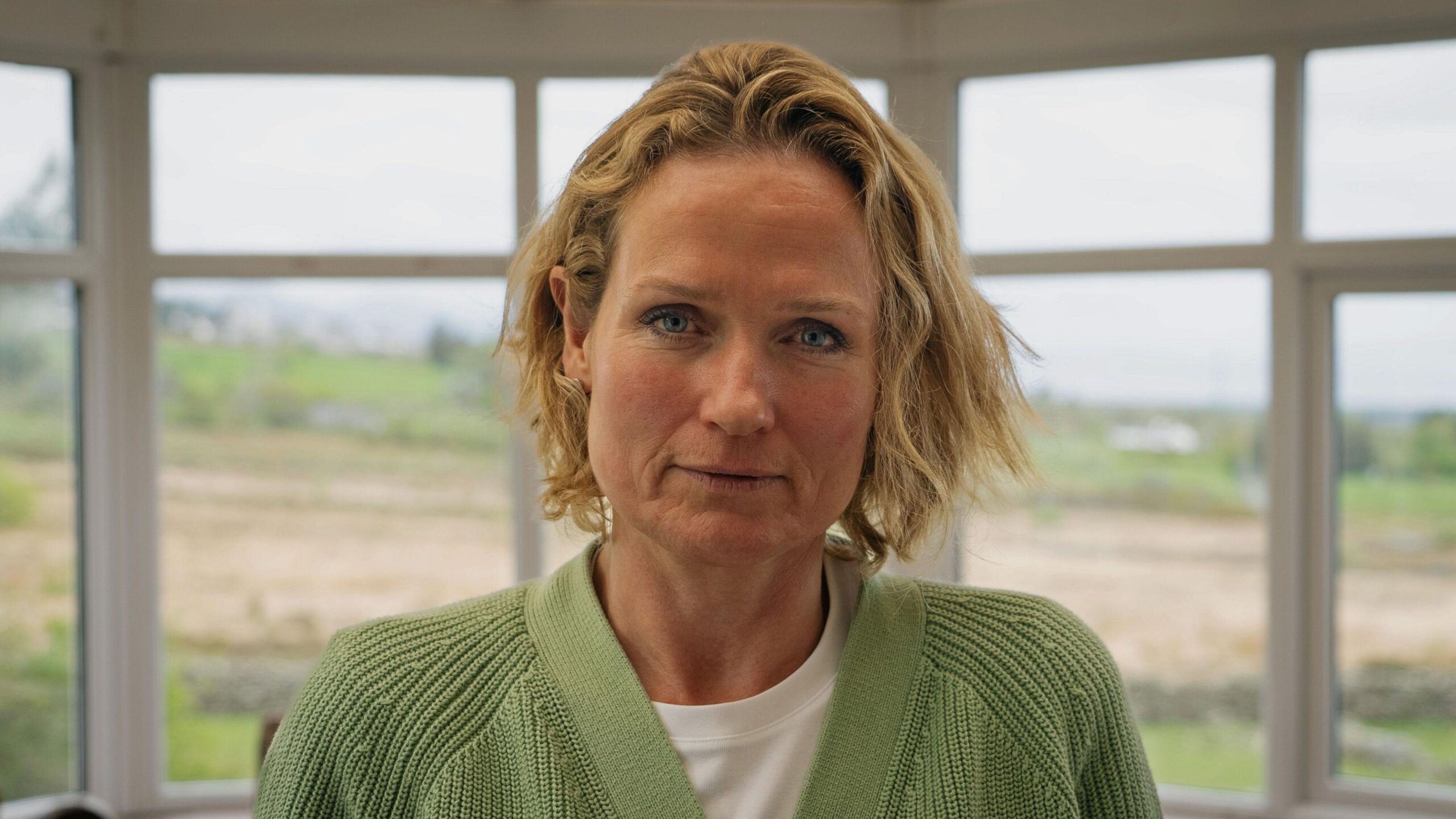 Rhianon Bragg looking at the camera. She has blue eyes and blonde hair, and has a neutral expression. She is wearing a green cardigan over a white t-shirt. She is standing in front of a window during the daytime.