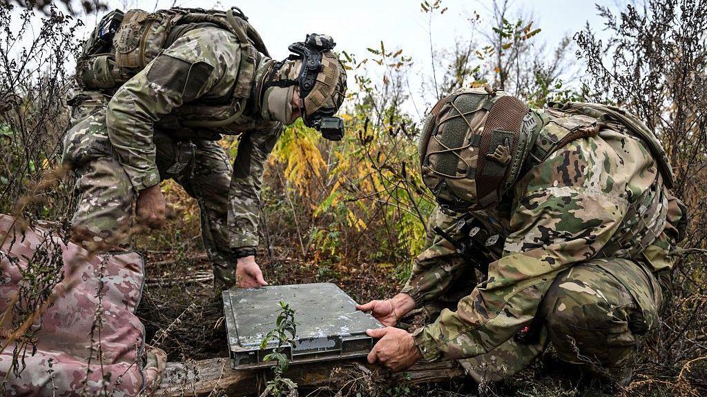 Two soldiers in green camouflage set up a Starlink terminal, which is also covered in green camouflage.