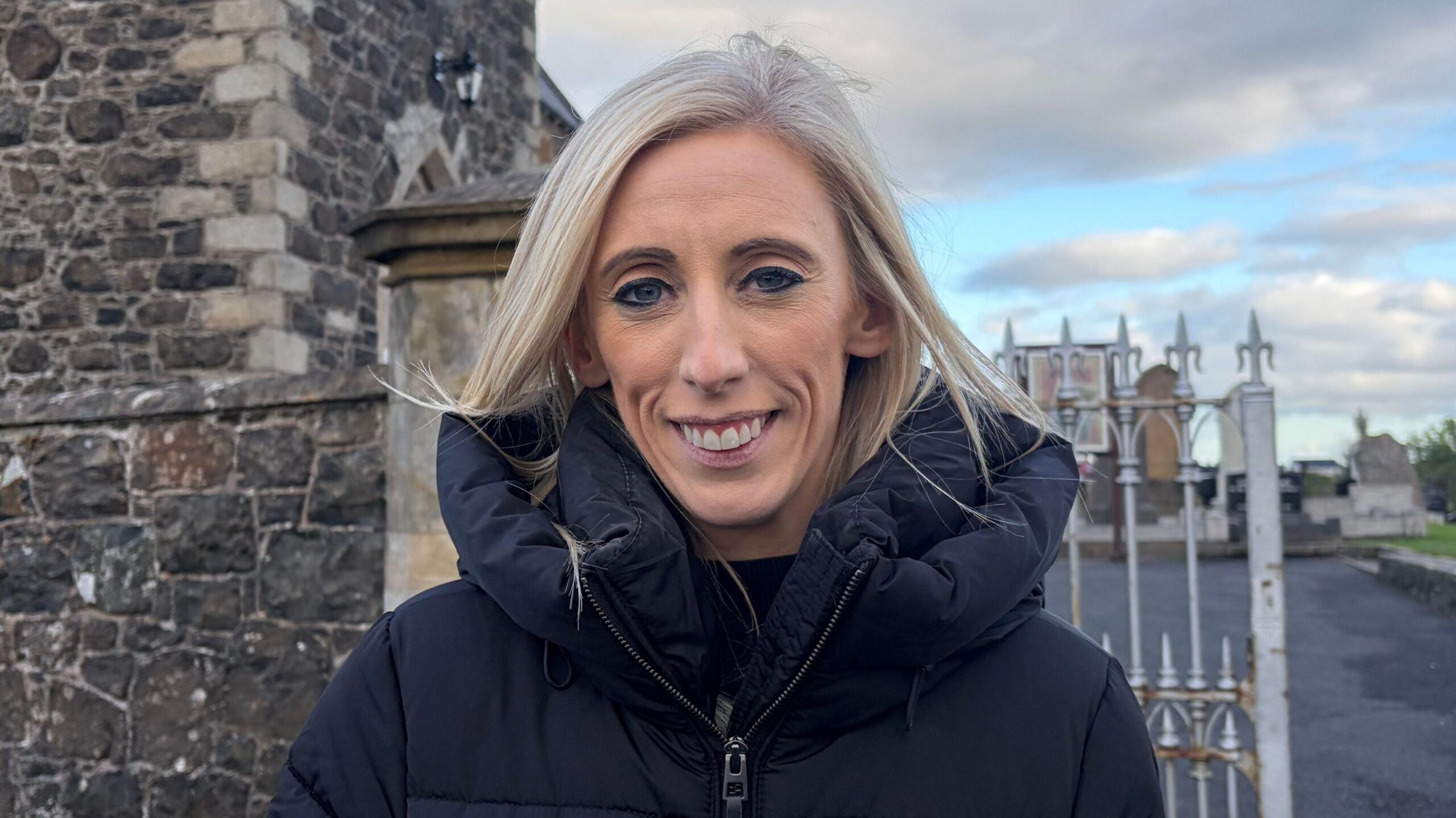 A woman with blonde hair smiles at the camera, she is wearing a black coat
