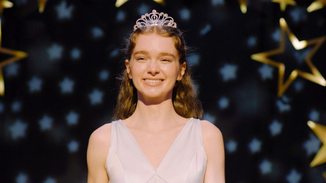 Stephen King’s Carrie returns to explore the horrors of social media A still from Carrie shows the main character smiling as she stands on a stage wearing a tiara and a white prom dress. Gold and white stars fill the background behind her, and a spotlight illuminates her.