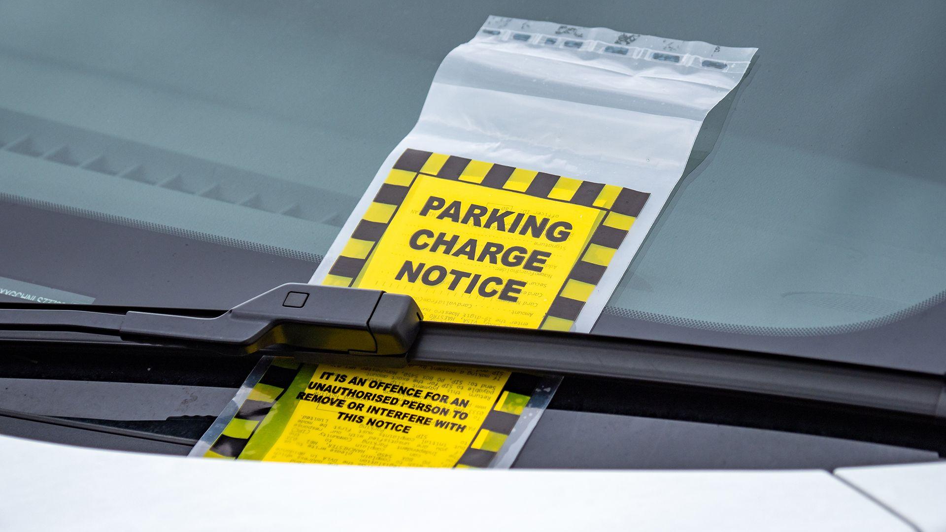 A yellow and black Parking Charge Notice under the window wiper of a car.