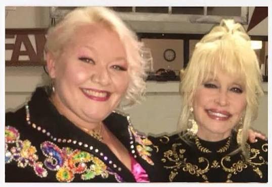 Jada Star and Dolly Parton are photographed standing next to one another. They both have blonde hair which is tied up. They are smiling and looking at the camera.