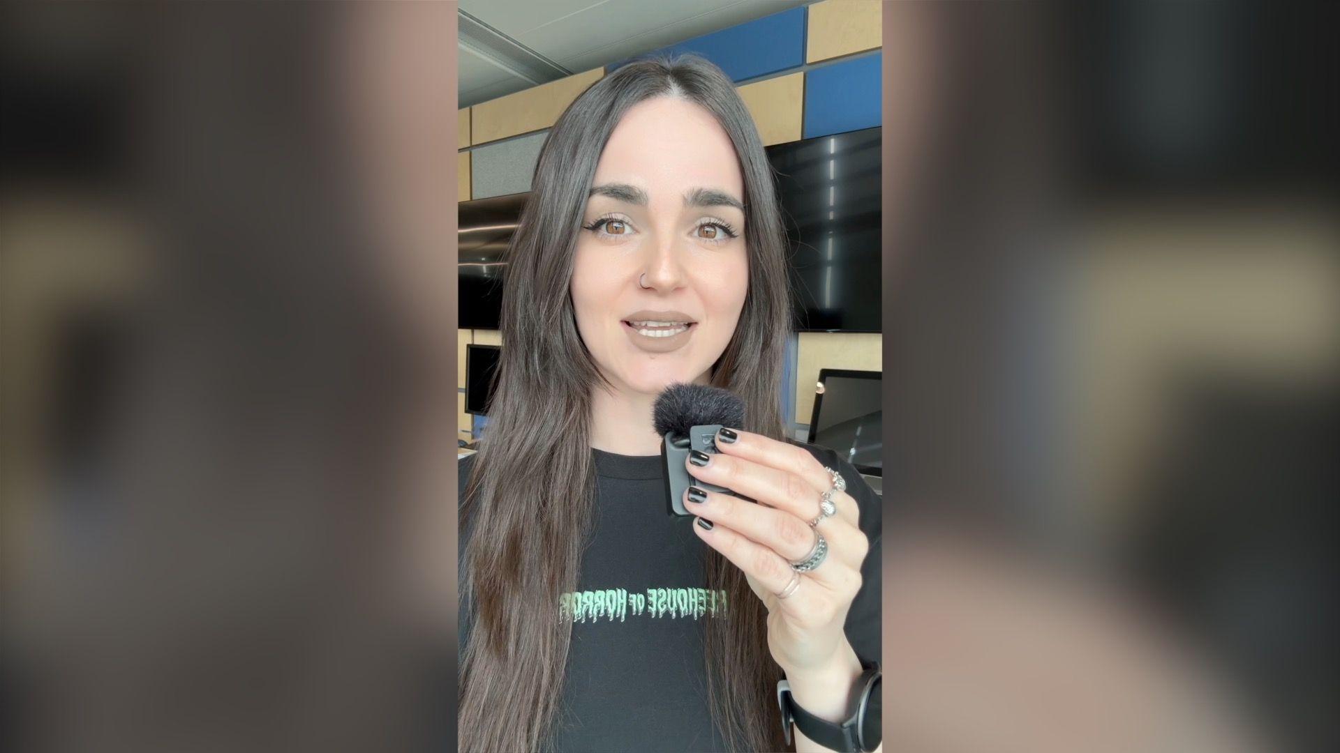 A woman with long dark hair, wearing a black T-shirt speaks into a clip-on mic.