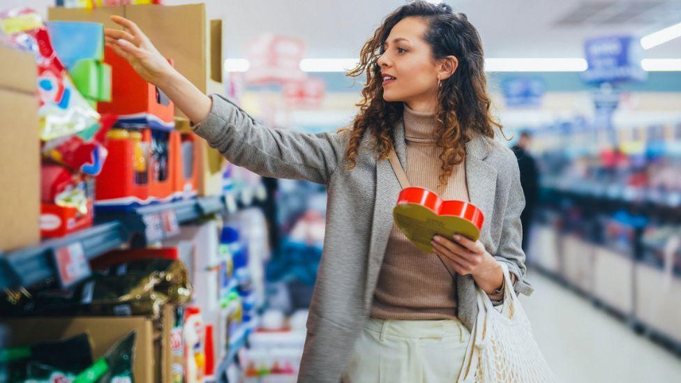 A woman compares items on a supermarket shelf. She has long, brown hair and is wearing a  grey jacket over a brown jumper and cream trousers.