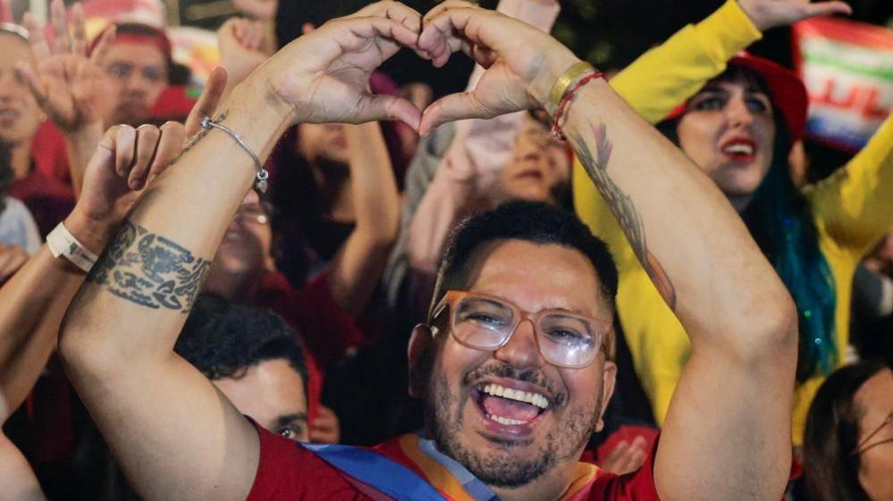 A bearded and bespectacled Lula supporter raises his arms to make the sign of a heart as he gives a broad smile. He is pictured in front of many other Lula supporters celebrating his victory.