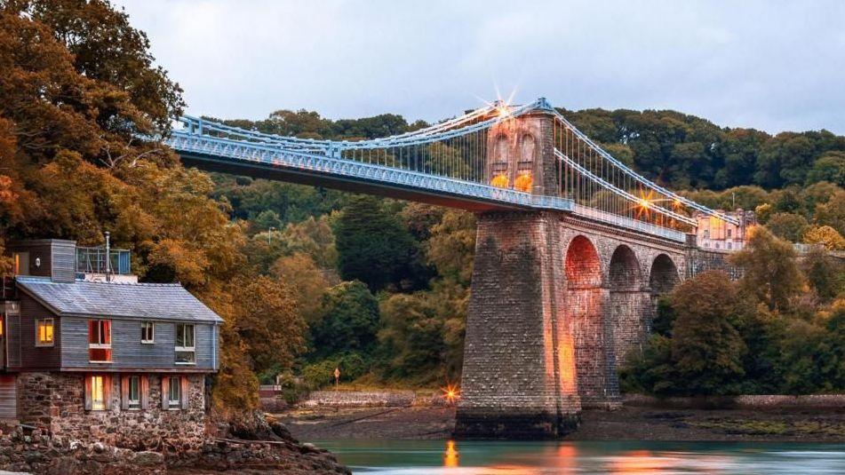 Menai Suspension Bridge, Anglesey, pictured from The Belgian Promenade. A house can be seen on the left hand side under the bridge. It is dawn and lights illuminate the bridge.