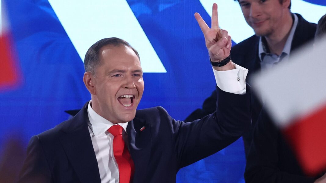 Police deployed during stand-off over control of Poland’s top court Karol Nawrocki stands on stage, gesturing the peace sign with his hand, wearing a dark suit, white shirt and red tie. A Polish flag is out of focus in the foreground.