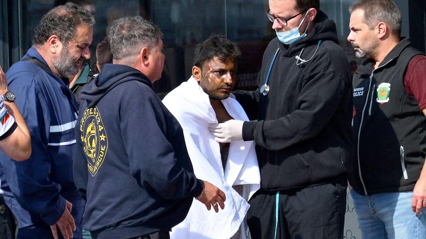 A man is wrapped in a large white blanket after being rescued from an attacked ship. There are people around him, one of whom has a stethoscope around his neck