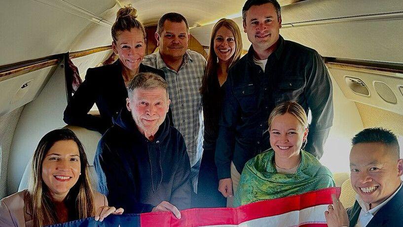Group crouched on inside of plane holding American flag. Hubbard in black hoodie with beard.