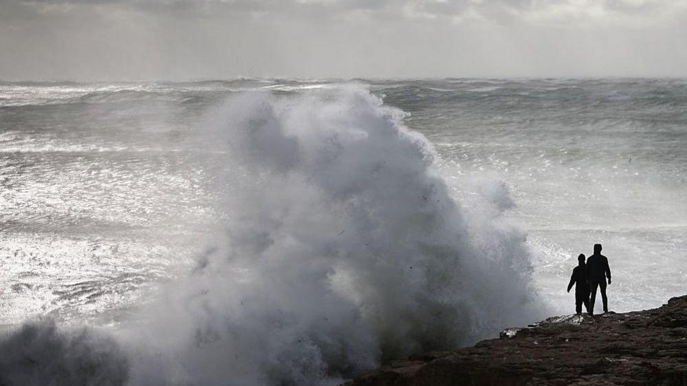 A large wave crashes against a rock jutting out to sea on the coast of England. The sky is grey. There are two people standing on the rock.