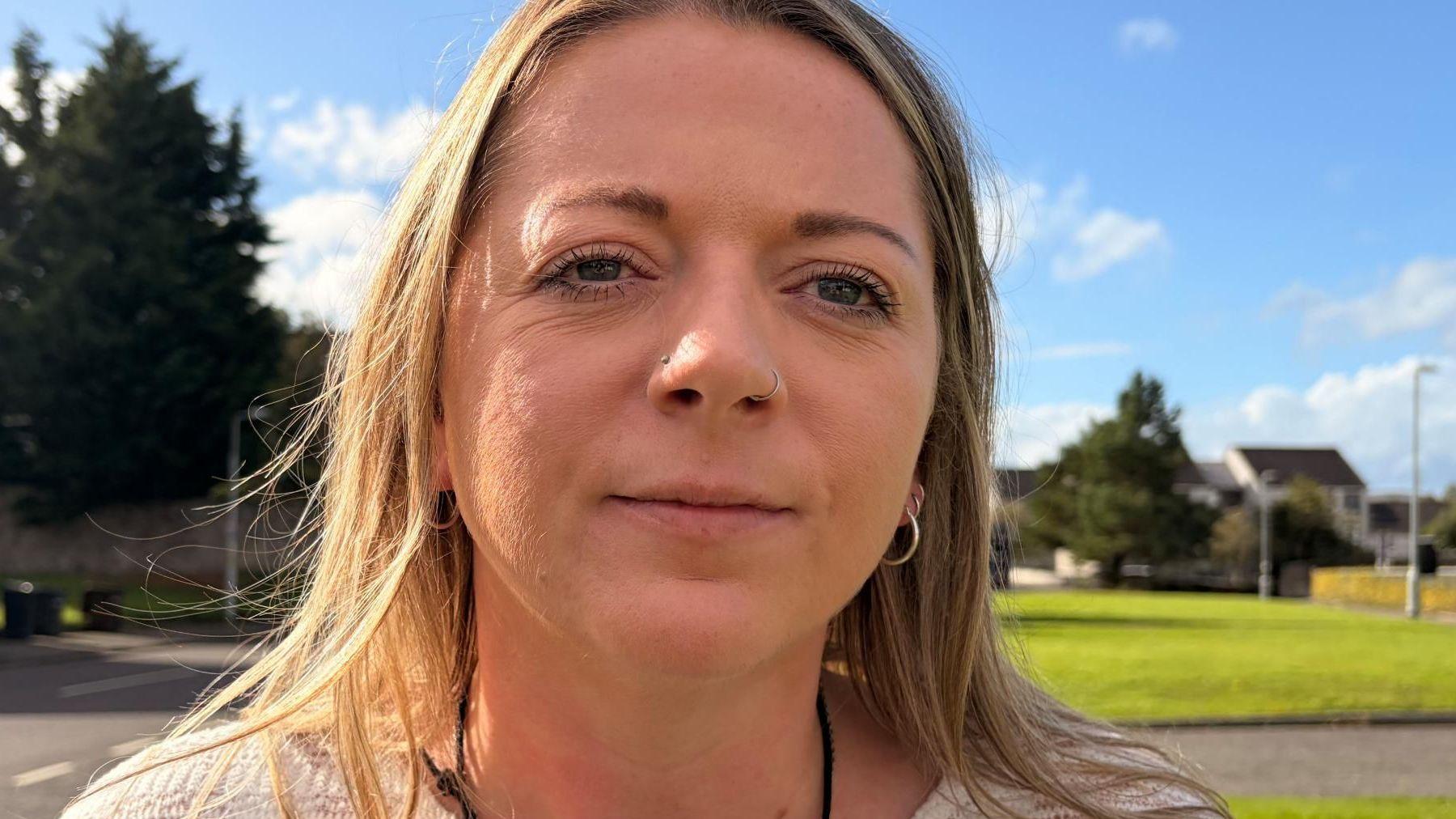 Close-up portrait of a woman with a nose ring and hoop earrings standing outdoors on a sunny day, with a grassy area, trees and houses blurred in the background.