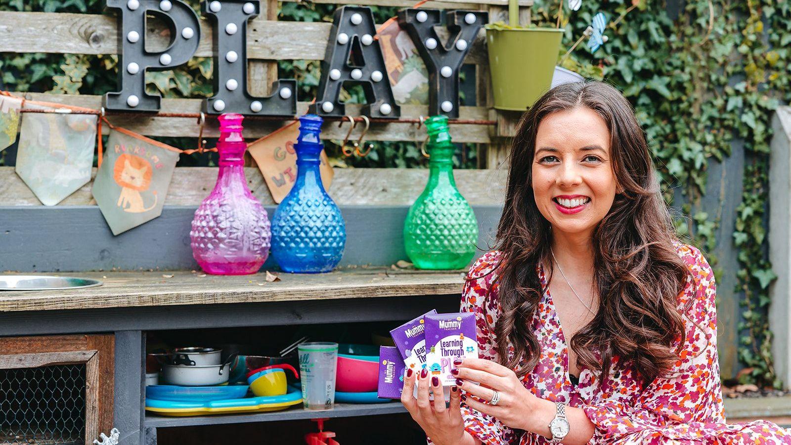 A woman with long dark hair, wearing a red, pink and white floral dress and a silver watch and ring, holds up three purple and white packets. Behind her is a wooden fence with ivy on it, a sign reading "play", some bunting and a surface with coloured bottles on it and plastic plates, pans and cups underneath.
