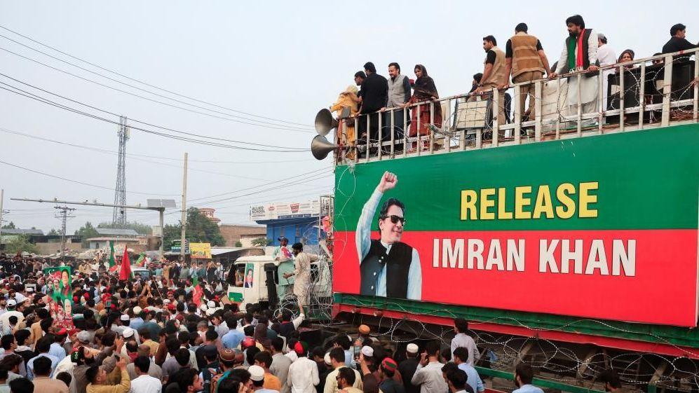 Dozens of men crowd together next to a large banner that shows Imran Khan with his fist pumping to the air. The banner is attached to a stage with more people standing on top