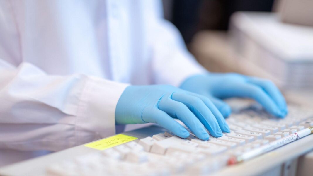 UK to invest £100m in ‘world first’ AI Biosecurity Lab A close-up stock image of a lab worker, wearing a white coat and blue gloves, typing on a keyboard.