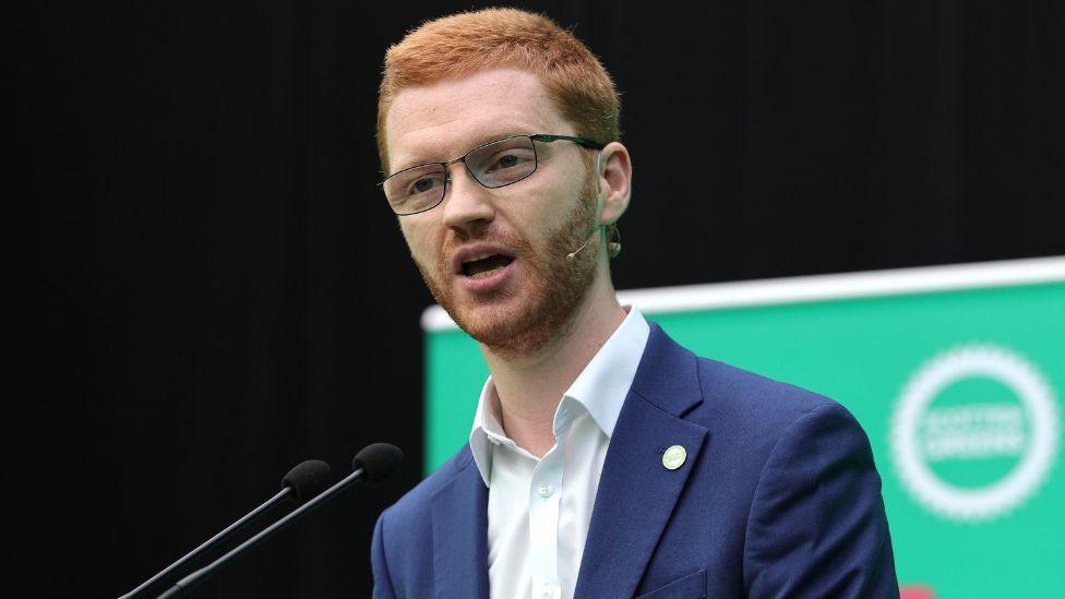 Ross Greer, who has short ginger hair, speaks into a microphone while wearing a business suit