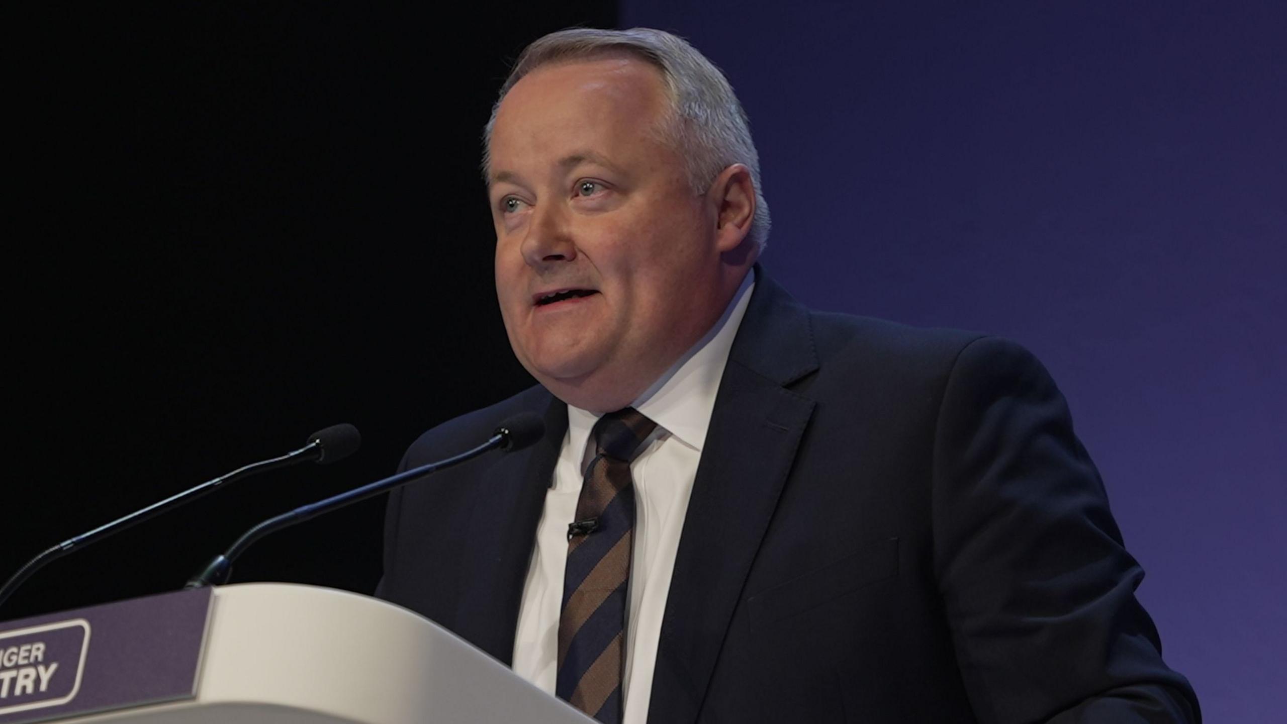 Darren Millar at a lecturn wearing a striped tie and navy suit jacket with a white shirt