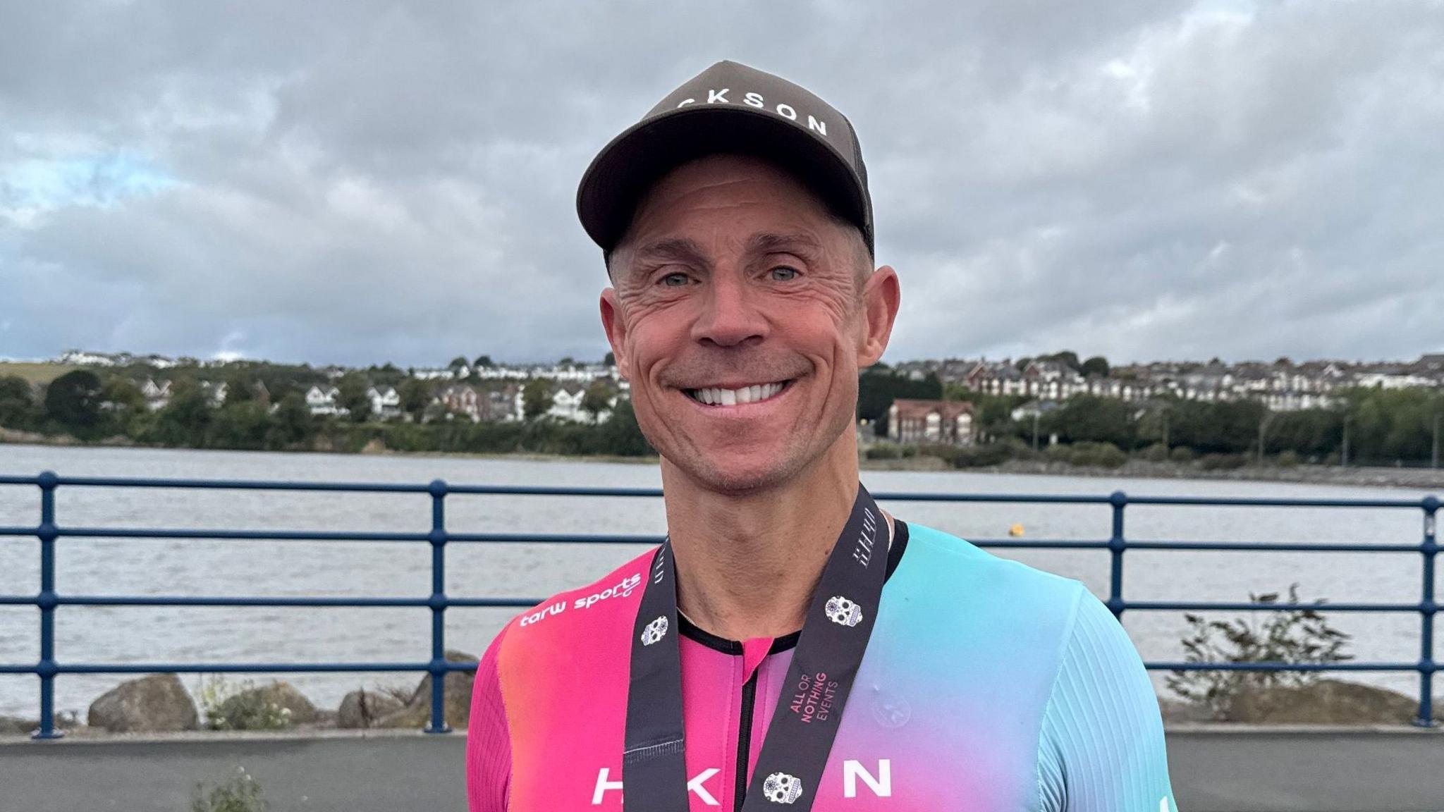 A man in a pink, blue and yellow triathlon suit and a medal around his neck. He's also wearing a cap and smiling at the camera.