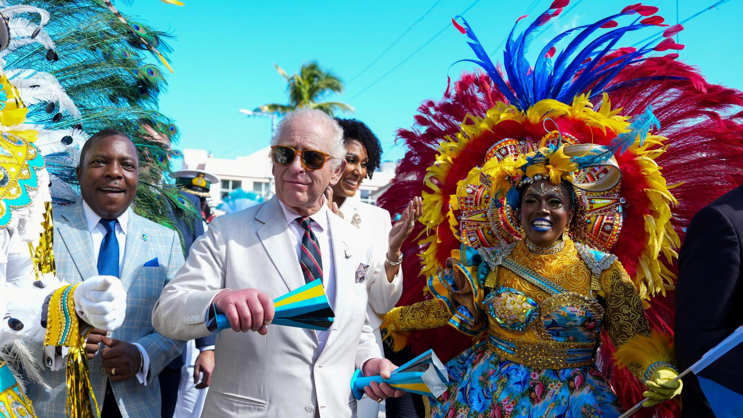 King Charles, wearing sunglasses and a grey suit, attends a Junkanoo parade and stands next to a man in a blue suit and a woman dressed in gold and blue parade attire.