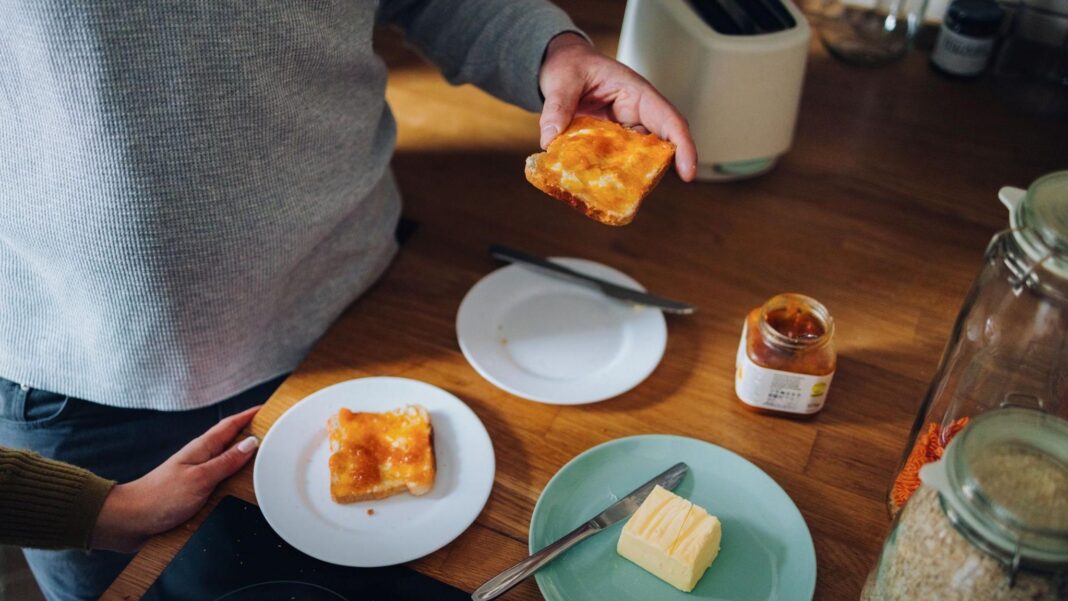 A man is visible from the chest down standing at a table eating marmalade on toast with a woman's hand visible in the shot.