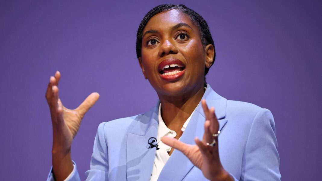 Kemi Badenoch speaking while wearing a light blue suit and white shirt. She's gesticulating with her hands as she speaks.